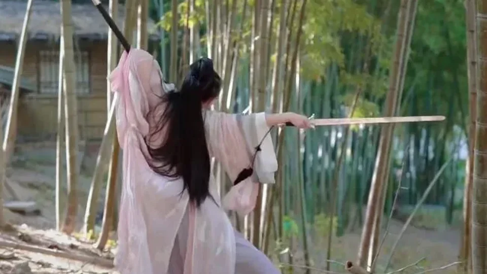 竹林里，一位身穿浅粉长袍的古装人物背向镜头持剑练习